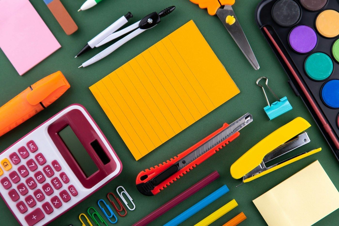 Office supplies including a calculator, stapler, notepads, and paper clips laid out on a green desk, representing bulk business purchases on Amazon Business
