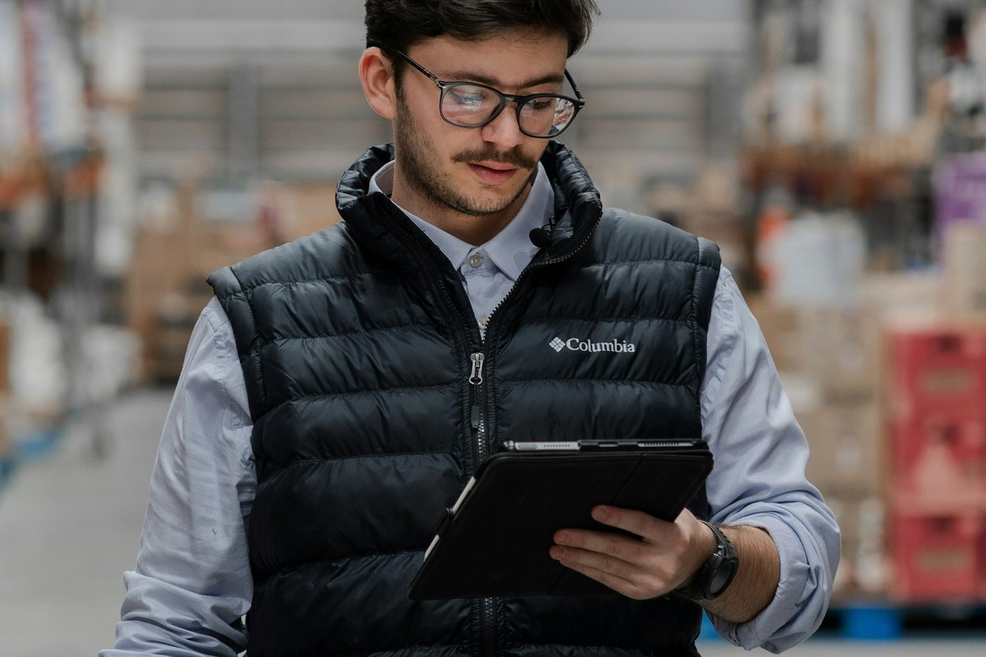 Warehouse worker in a vest checking inventory levels on a tablet in front of stocked metal shelves