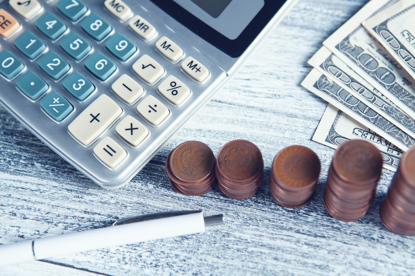 Calculator, stacks of coins, dollar bills, and a pen on a wooden table, representing checking your total price against the competitive external price in Pricing Health