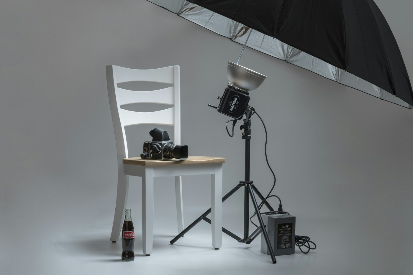 Photo studio with a camera on a white chair, a flash light on a tripod, and a large umbrella reflector, representing shooting new images for a main image experiment