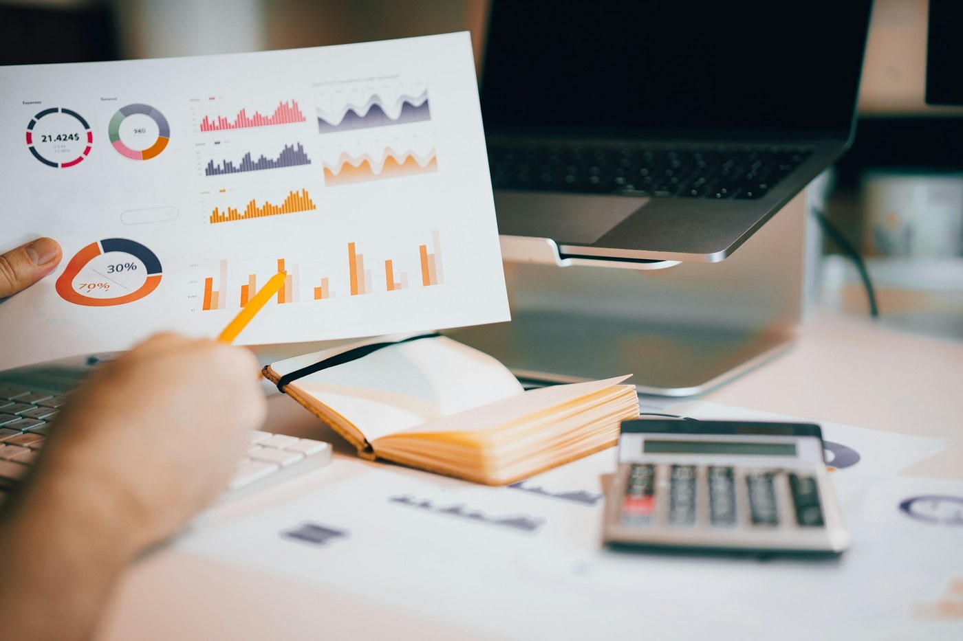 Hand holding a printed report of bar and donut charts above a laptop and calculator, representing reviewing niche metrics in Product Opportunity Explorer