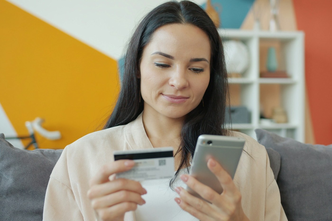 Smiling woman on a sofa holding a credit card and smartphone while shopping online, representing a repeat customer redeeming an Amazon Brand Tailored Promotion