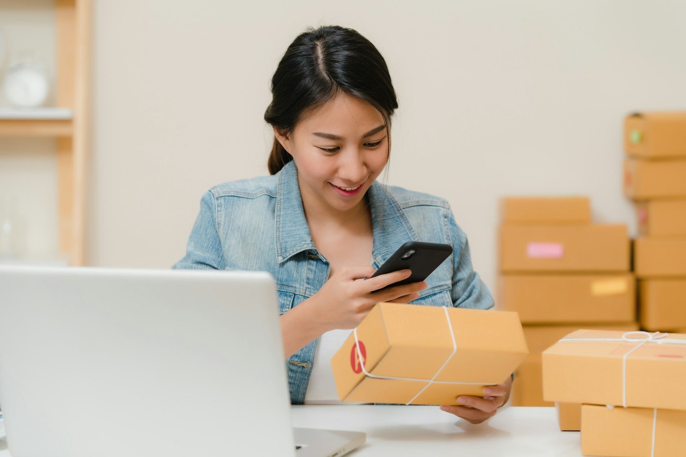 Seller at a laptop checking a packed shipping box with a phone, representing tracking and reducing Amazon FBA returns
